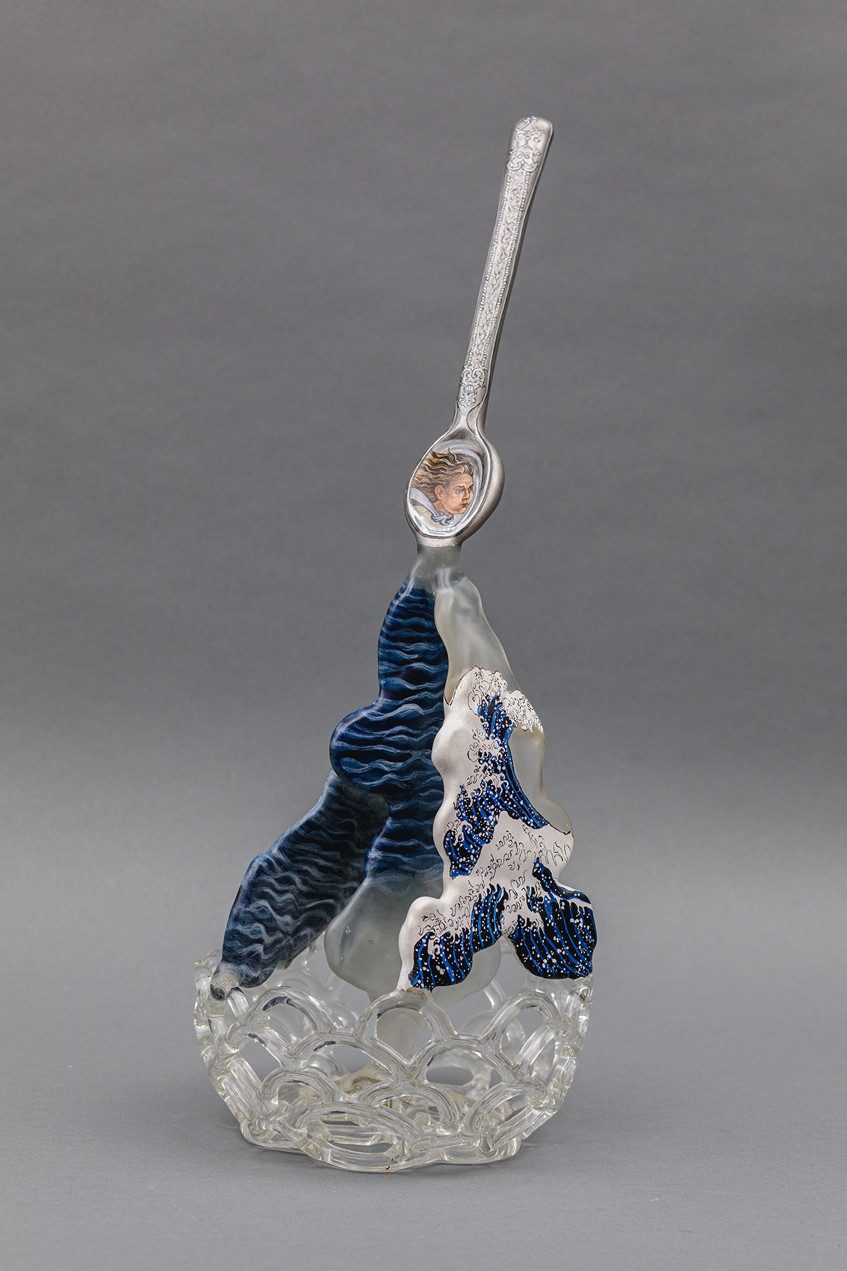 A spoon, holding the face of a person, is pouring an ocean with strong waves. The bottom of this piece is cup-like, built with many transparent arches.