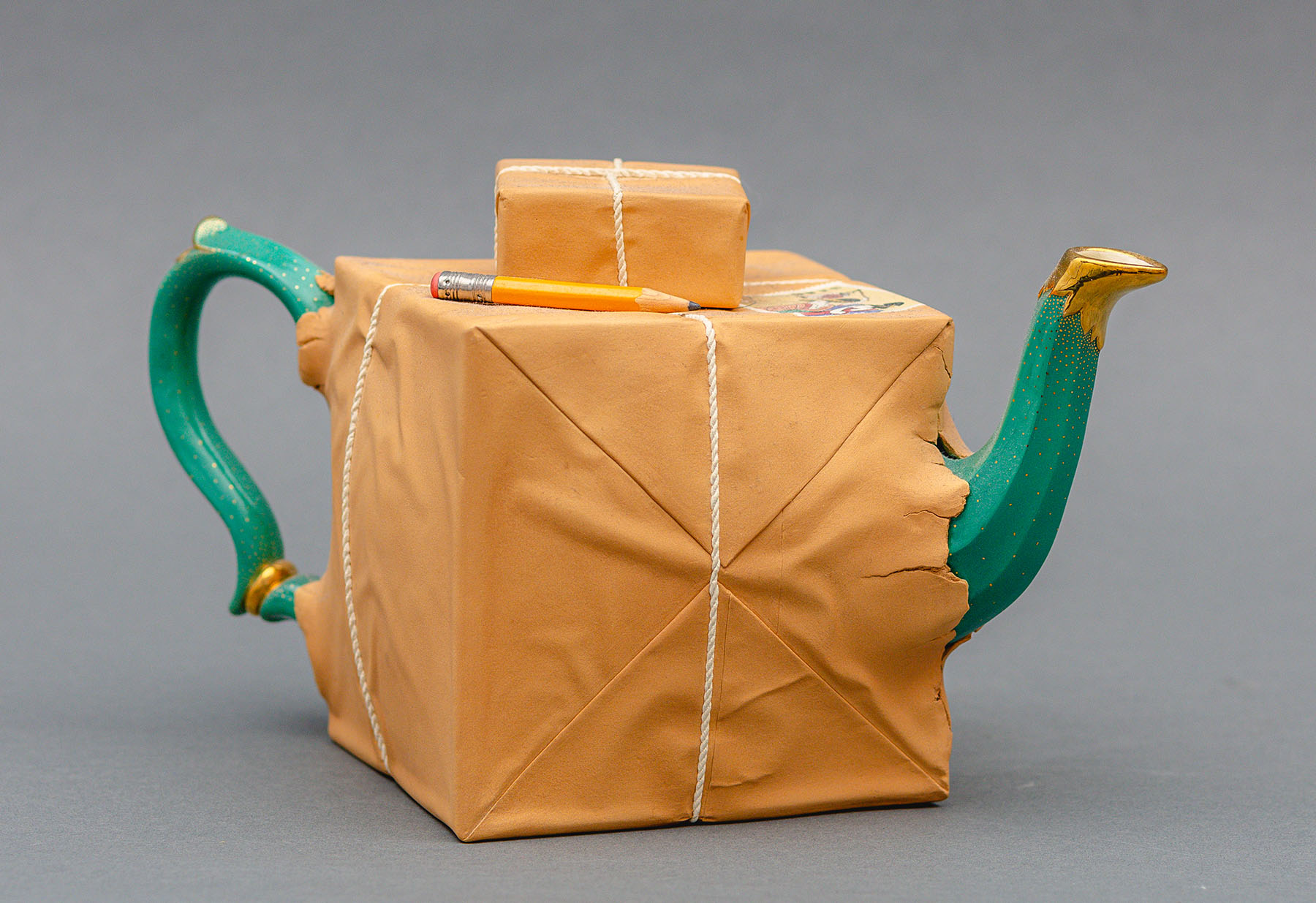 Karen Dahl, Box Too Small Teapot A ceramic teapot that appears as if the green and gold object is wrapped and sticking out of brown paper and rope. A smaller ceramic package and pencil sit atop the main portion.
