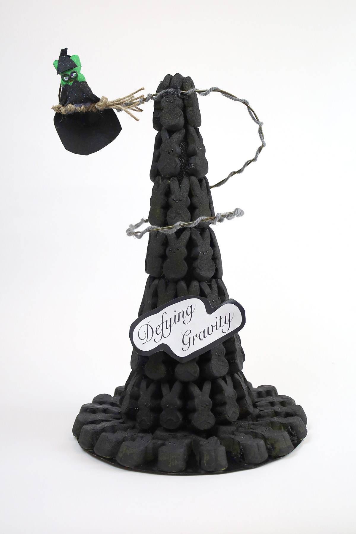 Kristina Tsioutsiopoulos Are Peeps Born Wicked A witch hat made from peeps and painted black, with a wire structure holding up a green peep riding a broom. There is text on the hat that reads "Defying Gravity".