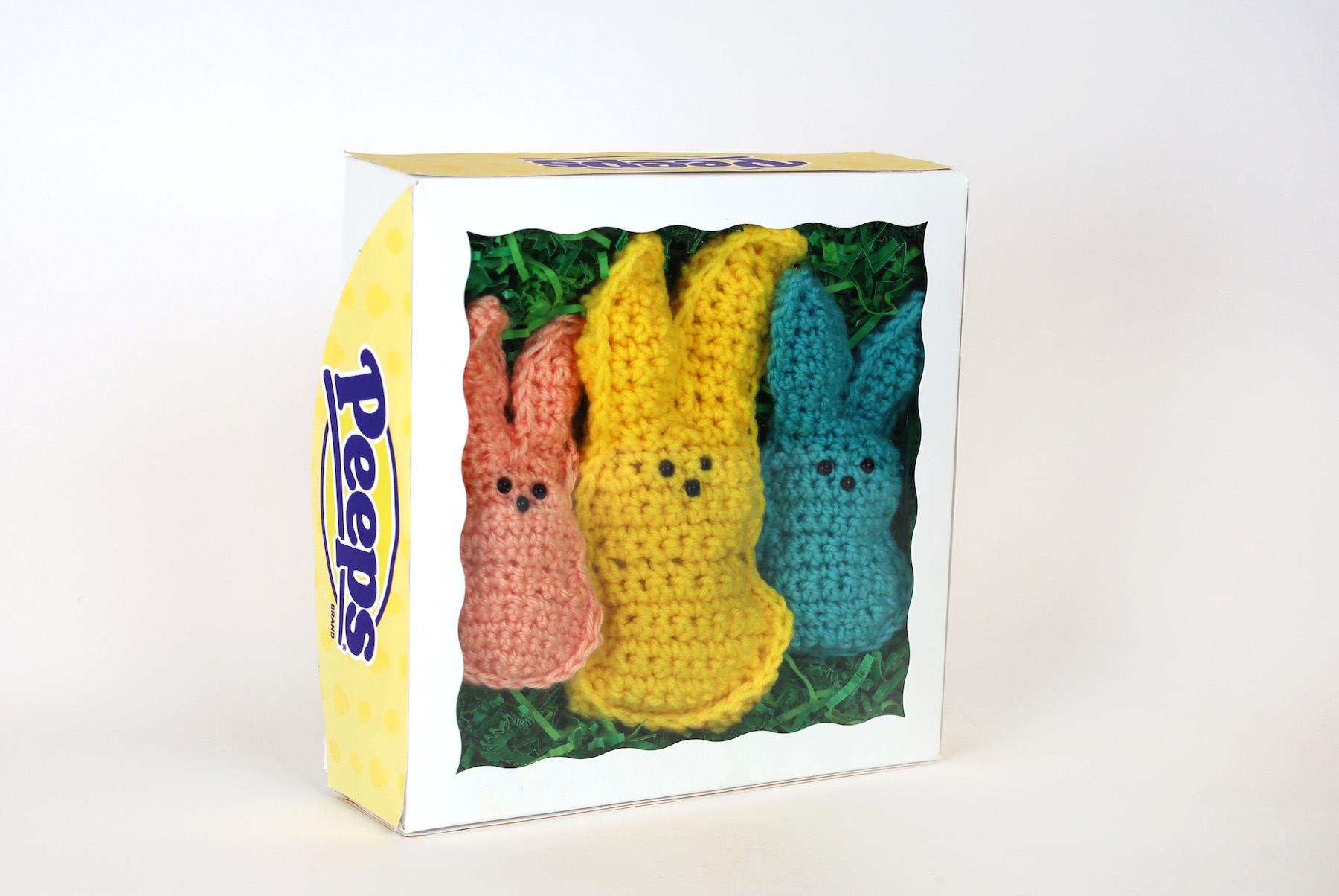 Azariah Niles, A Box Of Peeps Three knit bunny peeps are seen through the clear plastic of a peeps box. The bunnies are surrounded by shredded green paper that look like grass.