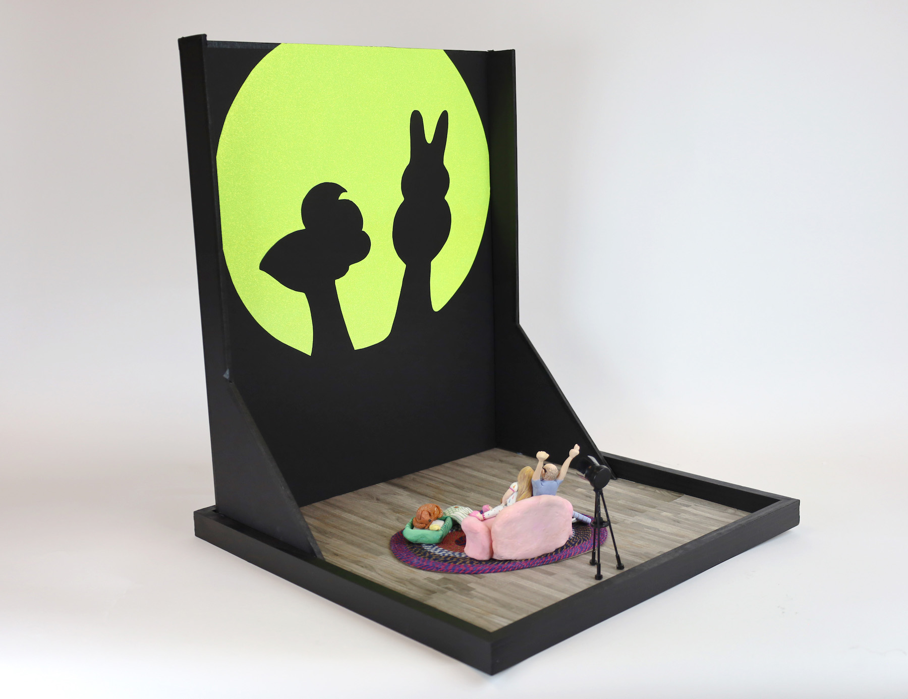 Beatrix Leach And Tom Spolar, Shadow Peepet Show (a) Two little clay people are on a couch with a dog in front of them. There is a spotlight behind the figure on the right. The figure on the right raises their hands to project a shadow puppet chick peep and a shadow puppet bunny peep on the wall.