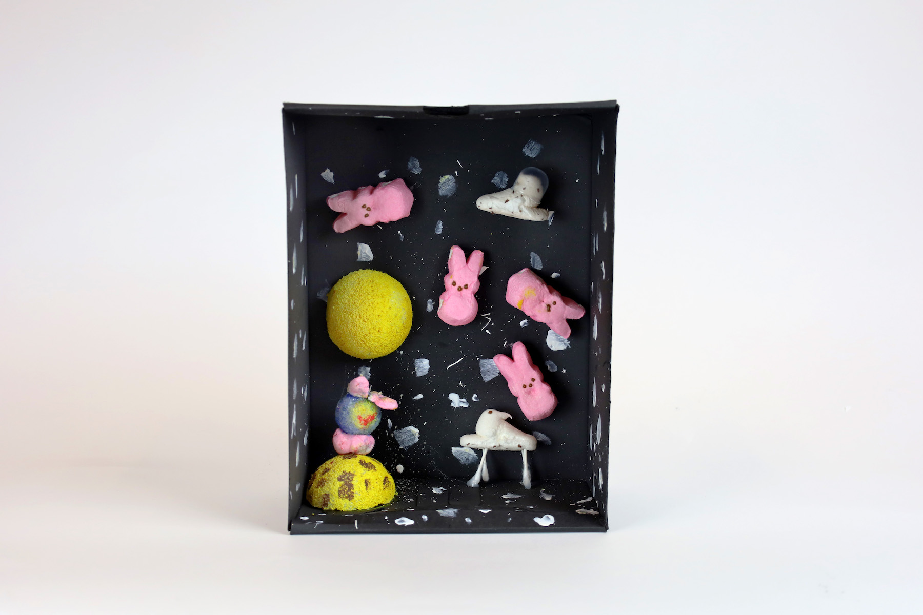 Maggie Weir, Peeps Go To Space! Peeps are suspended in a black box painted with white specks. There are two yellow spheres, one with brown spots, and a bunny peep whose head is replaced with a blue and yellow orb. This scene is of peeps floating through outer space.