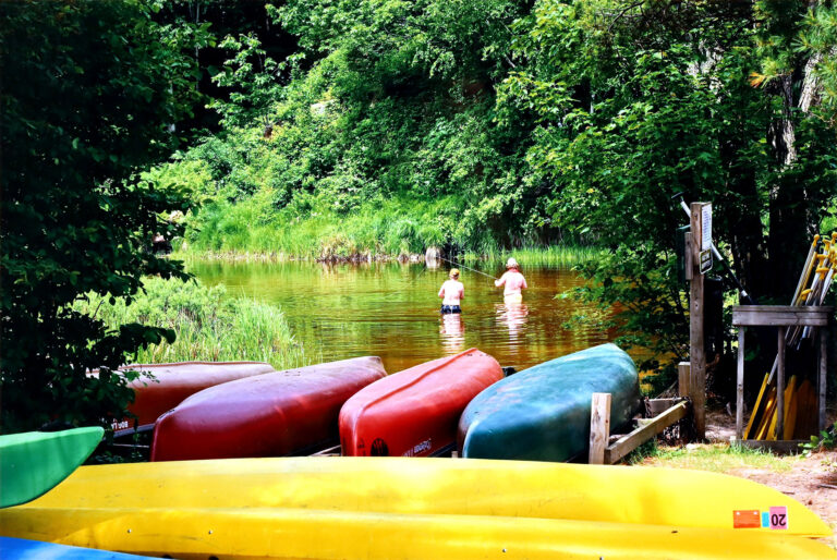 Photo of two people standing in water and fishing, surrounded by greenery. There are kayaks in the foreground.