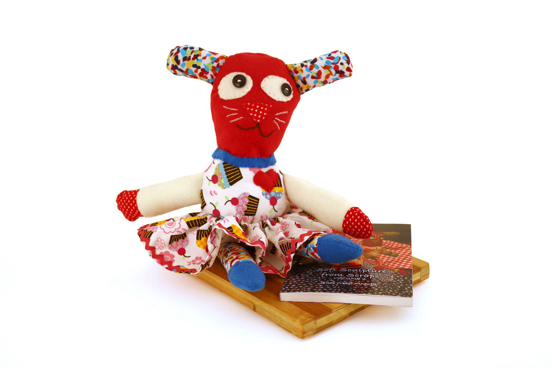 Janet Mrazek, Suzy-Q, I Love You! A stuffed animal with a dress and a red heart sits on top of a cutting board and a book. The book is called "Soft Sculpture from Scraps".