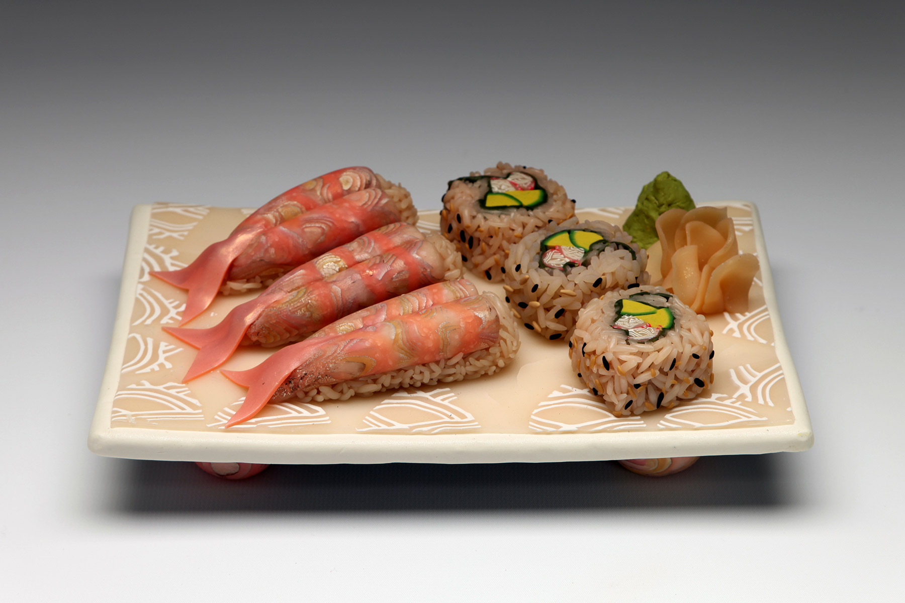 Lindly Haunani, Sushi Platter Polymer clay formed into the shape of a light tan/pink decorated platter. Atop the platter are three pieces of faux sashimi on the left with brown rice and pink fish; three pieces of faux sushi in the center with brown rice and green and pink filling; and faux ginger on the right with faux wasabi behind it.